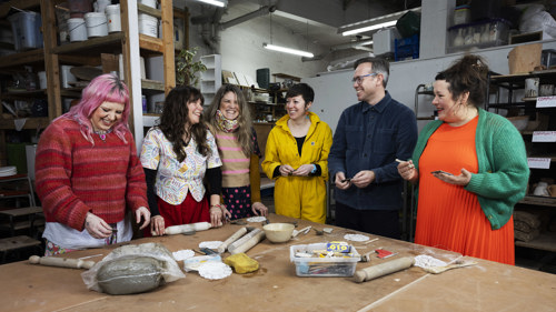 Artists from three collectives are at a table making buttons from clay, they are all dressed in brightly coloured clothes and laughing