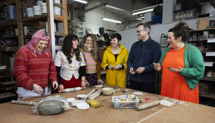 Artists from three collectives are at a table making buttons from clay, they are all dressed in brightly coloured clothes and laughing