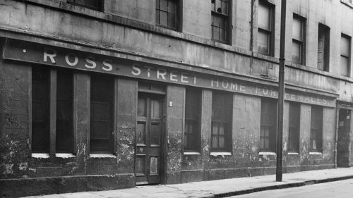 A black and white photograph of the exterior of Ross Street Homes for Females, in Calton. The title of the institution is written in capital letters along the front of the building. The walls are crumbling and the tenements above look dilapidated.