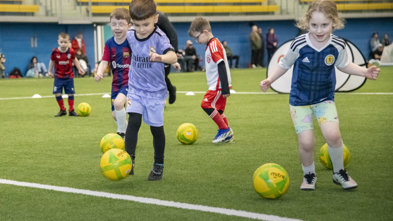 Young children in football tops dribbling yellow balls during an indoor football session on a green artificial surface