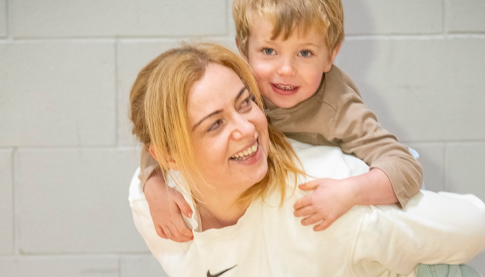 A smiling parent kneels on a mat indoors while a young child hugs their back, showing a playful moment during a parent-and-child fitness session.