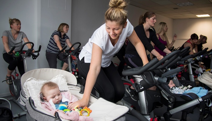 A group fitness class with a parent on an indoor bike whilst caring for their child in the pram. Combining indoor cycling and childcare in a supportive workout environment.