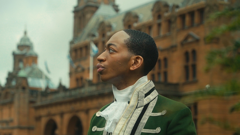 A young Black man dressed in 1700s style servants clothing, in the background is Kelvingrove Art Gallery and Museum