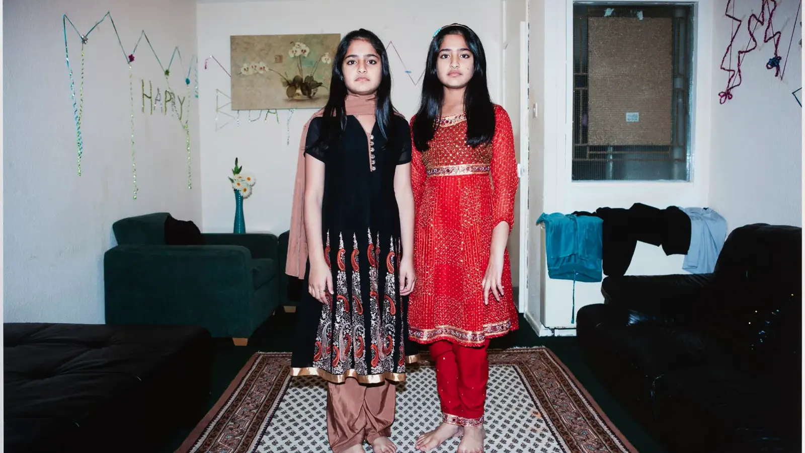 Two young girls stand side by side in a living room decorated for a birthday celebration. They wear traditional South Asian outfits—one in a black and brown patterned dress, the other in a red and gold ensemble—and are barefoot on a patterned rug. Behind them, “HAPPY BDAY” decorations hang on the wall above a green armchair, a black leather couch with clothes draped over it, and a small table holding a vase of flowers. 