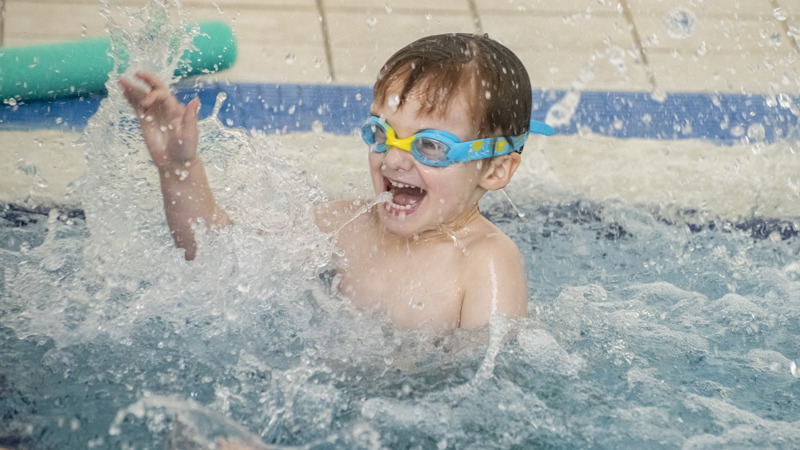 A young child splashing in a swimming pool they are smiling and wearing bright blue and yellow goggles