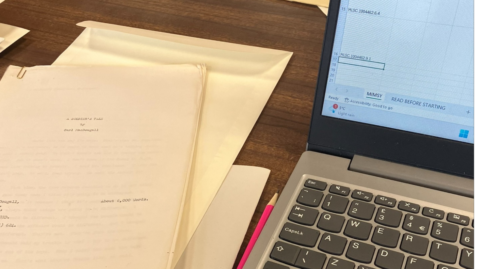 A wooden table in a library or study room is covered with typed manuscripts, folders, and a laptop. In the foreground lies a manuscript titled “A Soldier’s Tale” by Carl MacDougall, marked as about 6,000 words. The laptop screen shows a spreadsheet with accession numbers. A pencil rests between the laptop and the papers.