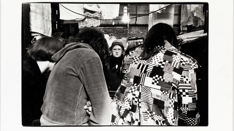 Black-and-white photo of a market stall with people browsing and patterned clothing in the foreground.