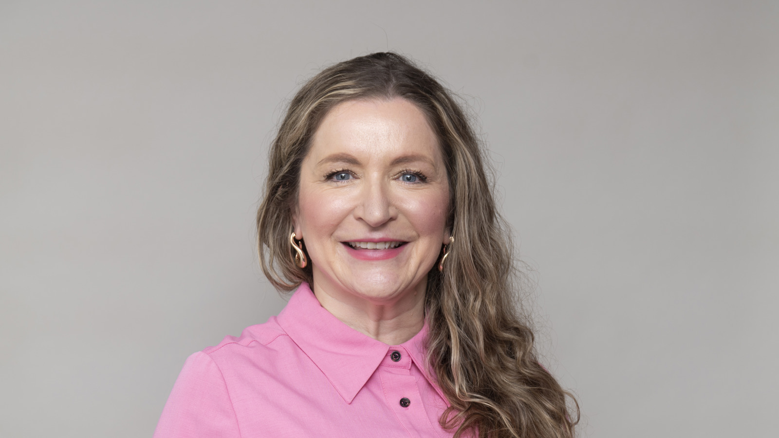 Headshot of a person with long wavy hair, wearing a bright pink shirt, smiling, against a neutral background.