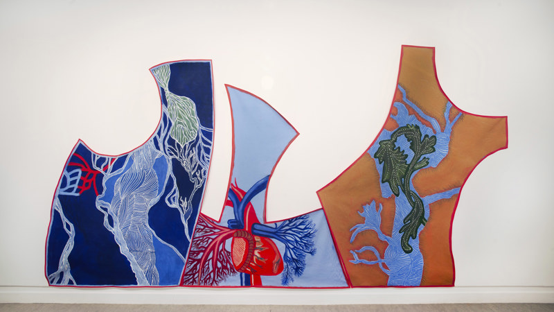 A large wall mural made of three irregular, abstract acrylic paintings on canvas, bias bound in red ribbon . The left panel is dark blue with branching white, green, and red vein‑like lines in a muscular form. The centre panel shows a detailed anatomical heart with red and blue vessels against a pale blue background. The right panel is a warm ochre coloured background  with a blue shape with a green acanthus leaf painted on top of it. The overall style is bold, colourful, and anatomical‑botanical in theme.