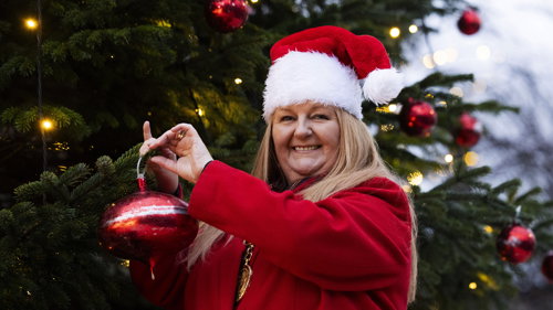 A person wearing a Santa hat and a bright red coat hanging a large red ornament on a Christmas tree decorated with lights and baubles.