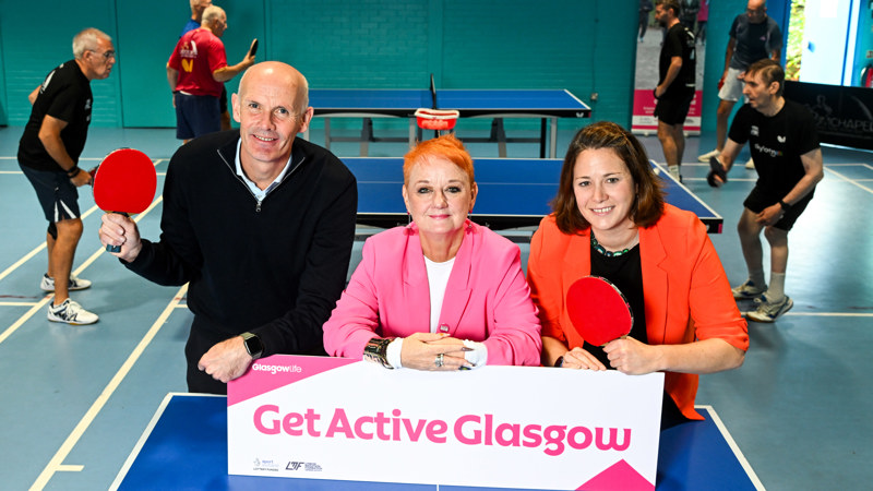A man and woman holding table tennis bats stand either side of a woman wearing a pink jacket leaning on a sign which reads 'Get Active Glasgow'
