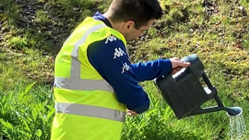 Person watering ground with watering can wearing a his vis jacket