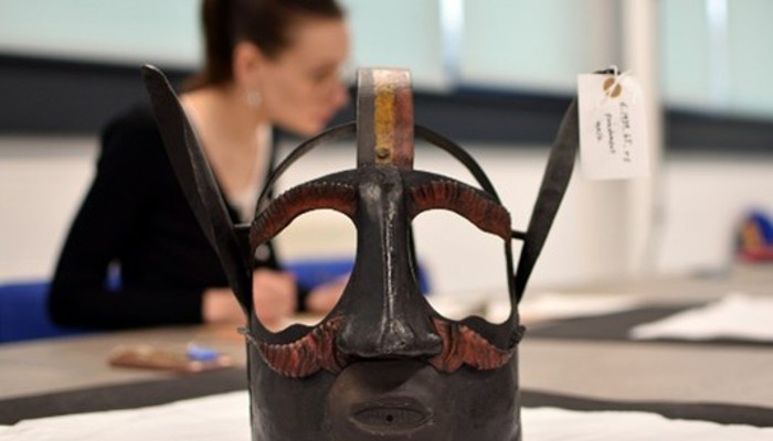 Photograph of a museum object with a blurred person in the background. The object is a mask made of metal designed to go over someone's head. It has ears, eyebrows and a moustache. The object label is attached to one of the ears and notes that it is a punishment mask.