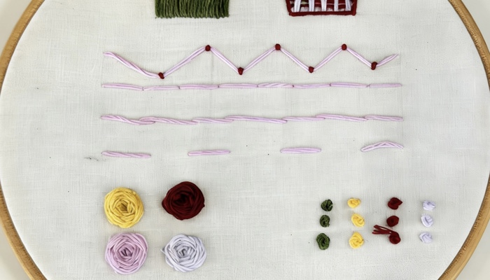 An embroidery sampler displayed in a wooden hoop, showing a variety of stitches on white fabric. The top sections include pink and green vertical stitches and a woven square in white and maroon. Below are rows of light‑purple linear stitches with small red knots. At the bottom are four circular floral motifs in yellow, maroon, purple, and lavender, with small grouped stitch samples in green, yellow, maroon, and lavender to the right.