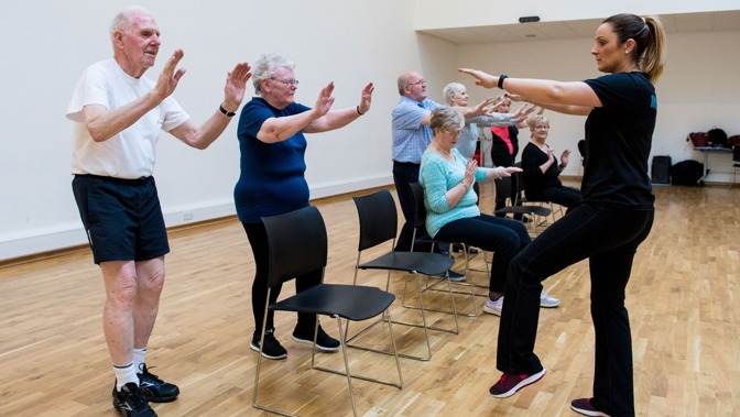 A fitness instructor taking a chair aerobics class with older participants, some are standing up while others are sitting down.