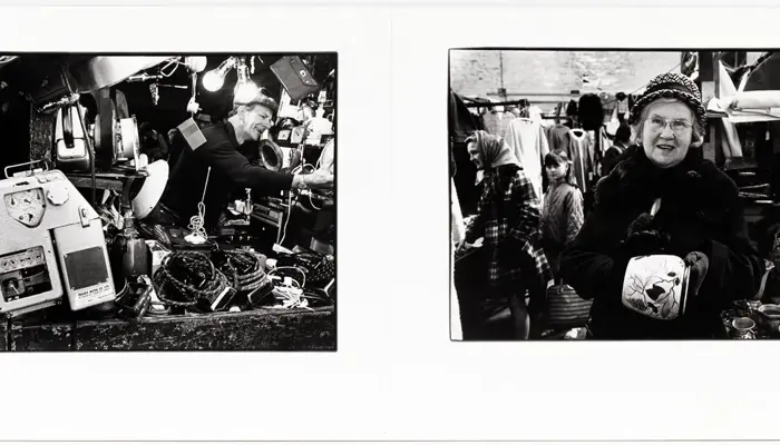 A black‑and‑white diptych shows two scenes from a busy outdoor market. The left image features a cluttered stall packed with vintage household items—ceramic dishes, kettles, a reel‑to‑reel tape recorder, and hanging lights—while a man reaches into the display, sorting through the goods. The right image shows an older woman in a patterned hat and fur‑trimmed coat holding a floral ceramic teapot in front of another stall filled with cups, textiles, and shoppers. 