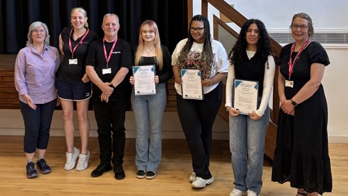 A group of seven people are gathered for a photo in a community centre. Three of them are holding certificates