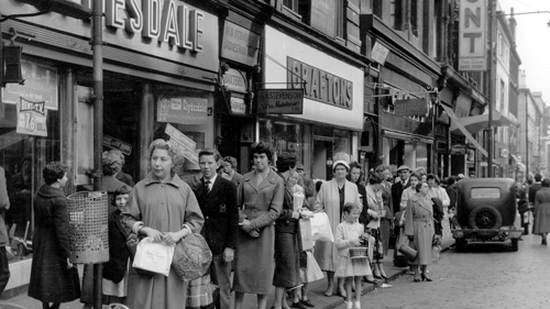 A black and white image of people lining in a queue for the bus on Sauchiehall Street from 1958