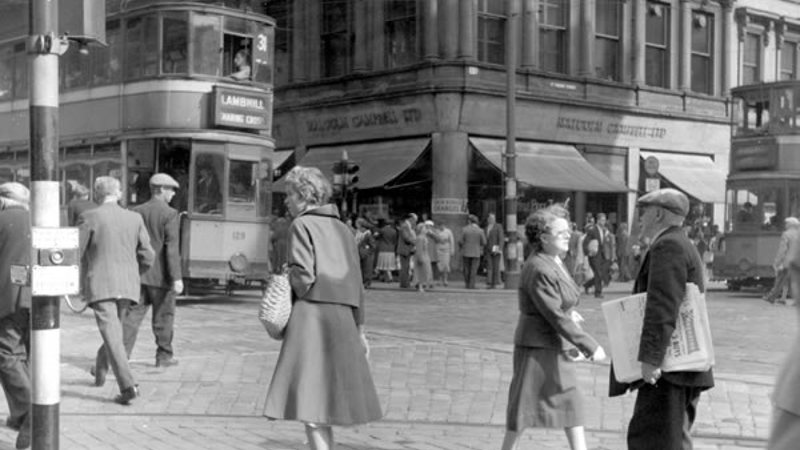 A black and white photo of a busy street with trams, many pedestrians, high sandstone shop buildings and traffic lights.