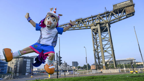A person in a colourful unicorn mascot suit is jumping in the air in front of a large industrial crane beside a river