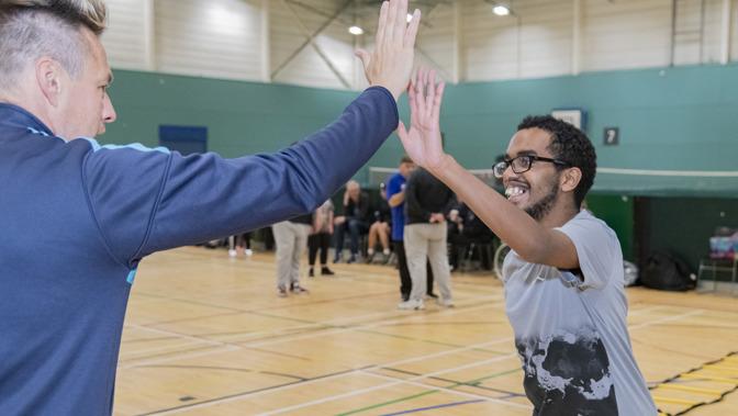 A coach and a young participant high-five on an indoor sports court, with other people and equipment visible in the background.