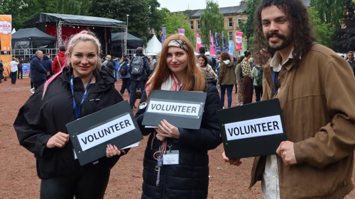 A group of three people holding clipboards that say volunteer on them at an outdoor musical event in a park