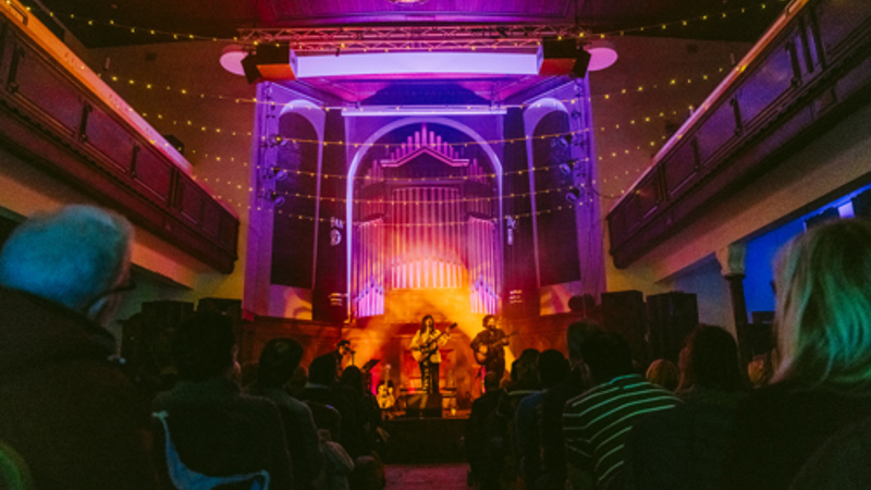 Audience watching a live music performance in a church-style venue illuminated with colourful stage lighting.