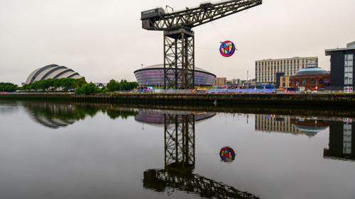 A large scale colourful rose shaped sculpture is hanging for a large industrial crane on a city riverside