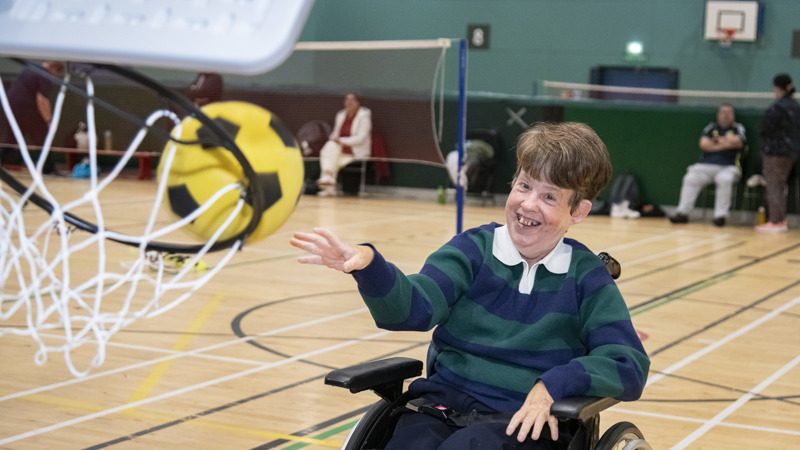 A person in a wheelchair smiles while throwing a large yellow and black ball near a basketball hoop.