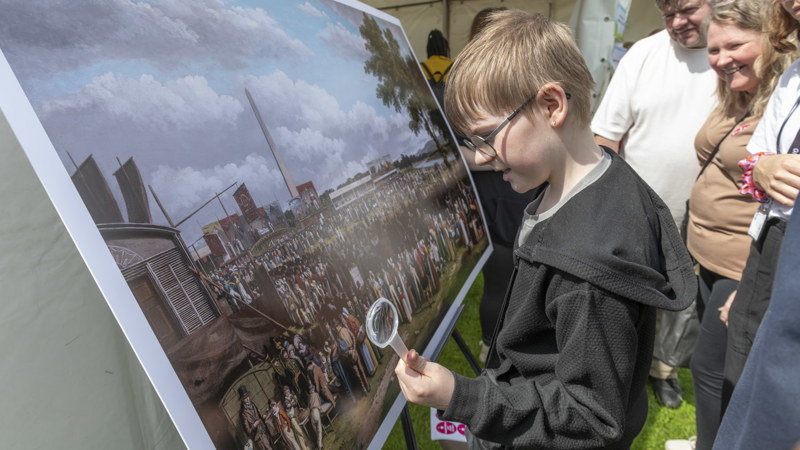 A child looking closely at a painting while holding a magnifying glass, other adults are standing behind them.