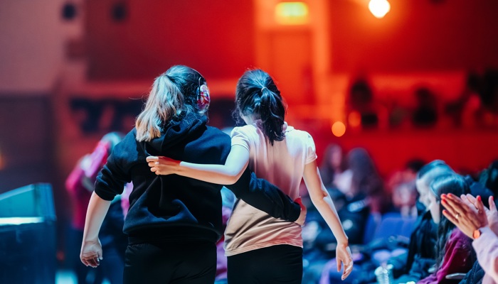 Two young girls with their arms around each other, watching a performance in a concert hall with blue and red light.