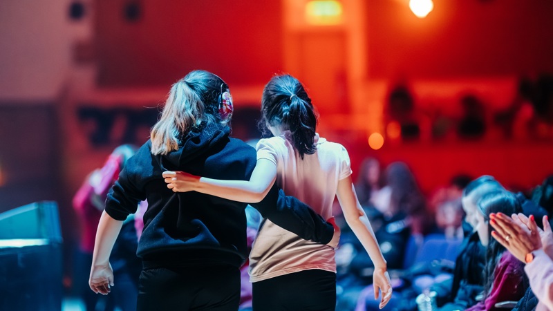 Two young girls with their arms around each other, watching a performance in a concert hall with blue and red light.