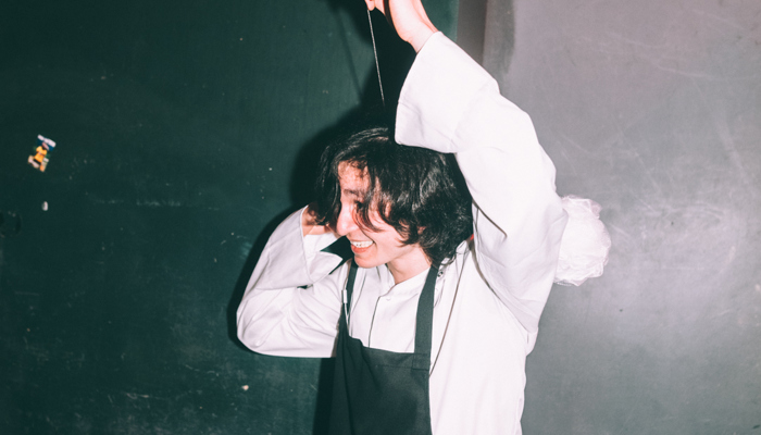 A person stood with their hand over their head, wearing cheffing whites and an apron 