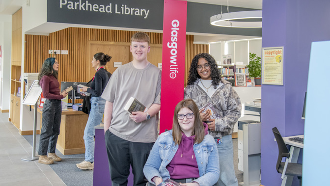 A group of three people holding books pose inside Parkhead Library, with one person seated in a wheelchair at the front. Two more people stand behind them. In the background, two people speak near an information desk, and bookshelves and computers are visible throughout the bright, modern library space. A tall sign reading “Glasgow Life” stands beside the group.