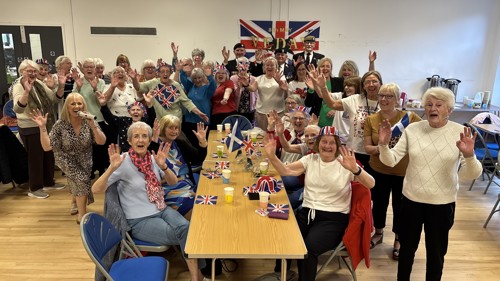 A large group of people cheering at a VE Day celebration event in a community centre. Some people are sitting at long tables, others are standing behind with their arms in the air. There are Union Jack flags decorating the hall.