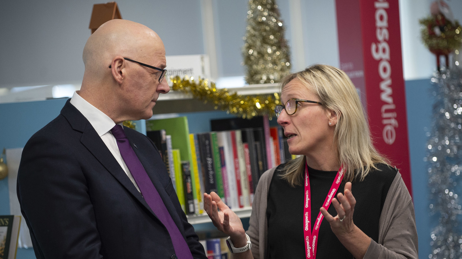 A man and a woman talk to one another in front of a book shelf which is decorated with gold tinsel and a small Christmas tree