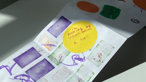 A drawing is on a table on it is prints and writing, it is brightly coloured with yellow and purple. 