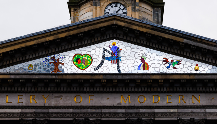 A neoclassical building’s pediment decorated with a colorful mosaic featuring symbolic figures, shapes, and animals sits above a stone façade with gold lettering. Above the pediment, a clock tower rises with a round clock face showing the time around 10:10.