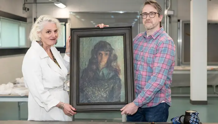 Two people stand side by side in a bright gallery-like room, holding a framed portrait between them. The woman on the left wears a white coat, and the man on the right wears glasses and a pink-and-blue plaid shirt. In front of them, a table displays several decorative objects, including a patterned ceramic pitcher, a matching bowl, a necklace, and a folded textile. The scene suggests an art or museum setting focused on curation or preservation.