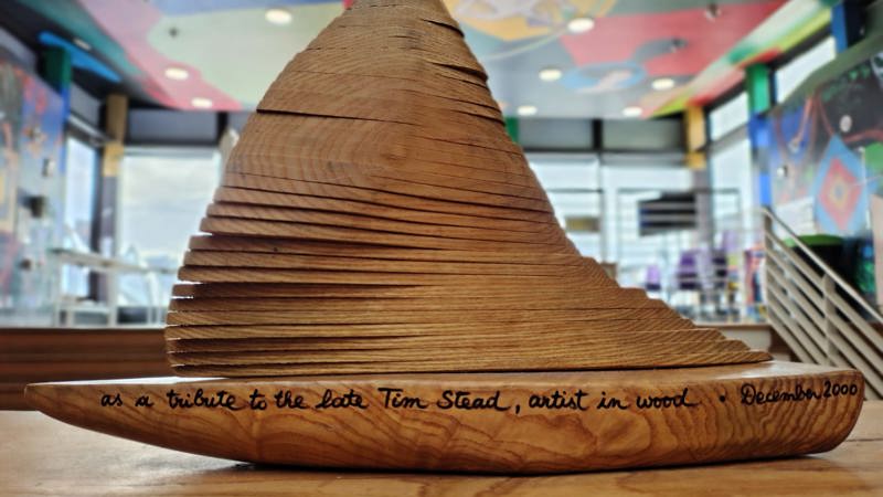 A wooden sculpture shaped like a boat, with an inscription reading "as a tribute to the late Tim Stead, artist in wood. December 2000"