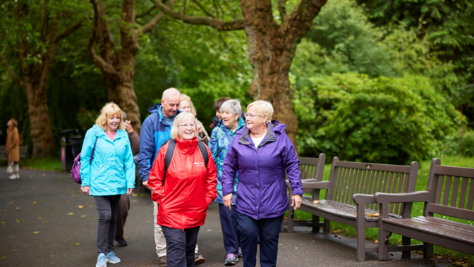 A group of older people chatting and walking together through a leafy park with wooden benches in the background.