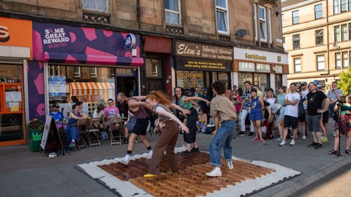 A group of four dancers are performing for a crowd on a piece of parquet flooring they have placed in the middle of a shopping street 