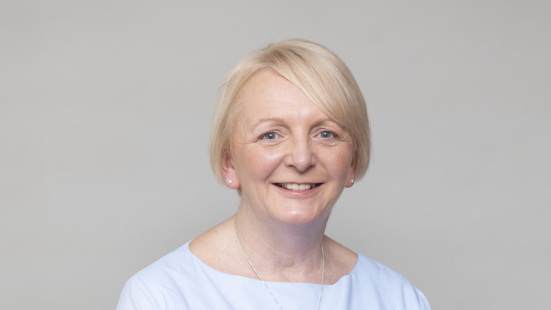 Headshot of a person with short light hair in a pale blue top, smiling, against a neutral studio background.