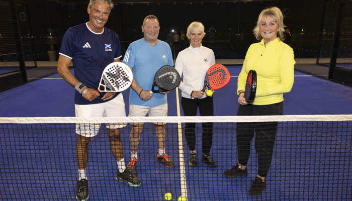 Four padel players stand behind the net while holding racquets on the same side of a padel court