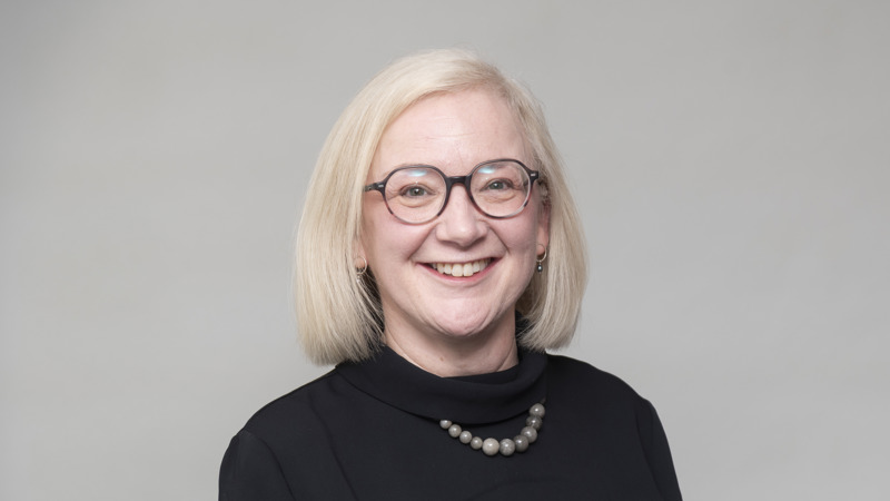 Headshot of a person with light hair and glasses, wearing a dark top and necklace, against a neutral background.