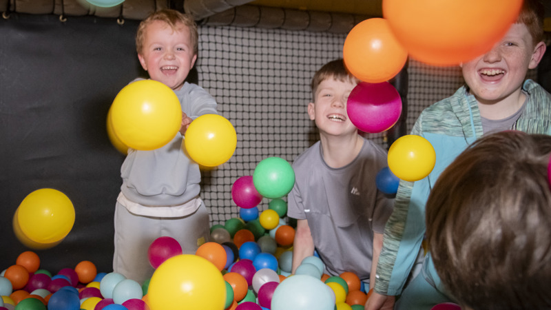 Children are laughing and playing a ball pit, a boy is throwing multiple balls in the air and smiling at the camera