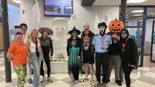 A group of nine people dressed up for Halloween in a sports centre reception area. There is a witch, a pumpkin, a wrester and a ghost.
