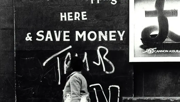 A black‑and‑white photograph shows a woman walking along a sidewalk past a wall covered in bold painted advertisements and a partially torn poster. The wall features large block lettering promoting a grain store and urging shoppers to “save money.” Beside it, a poster about inflation shows a candle shaped like a currency symbol burning at the top. The woman wears a coat and carries a bag, moving through an urban street setting.