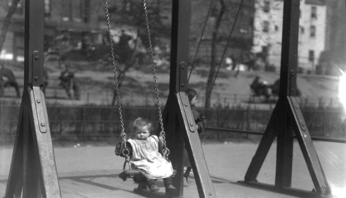 A black and white photograph of a baby on a swing holding onto the swing chain, wearing dirty clothes. There is an older child on the other swing and buildings in the background.