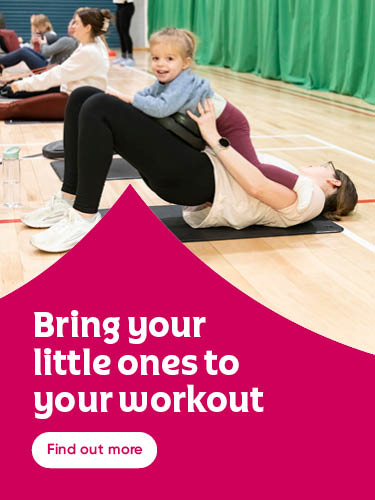 A woman lying on a gym mat performs a glute bridge exercise while holding a small child on her stomach. In the background, other adults participate in a group workout session on a wooden gym floor. The image includes the text "Bring your little ones to your workout" and a button that says "Find out more."
